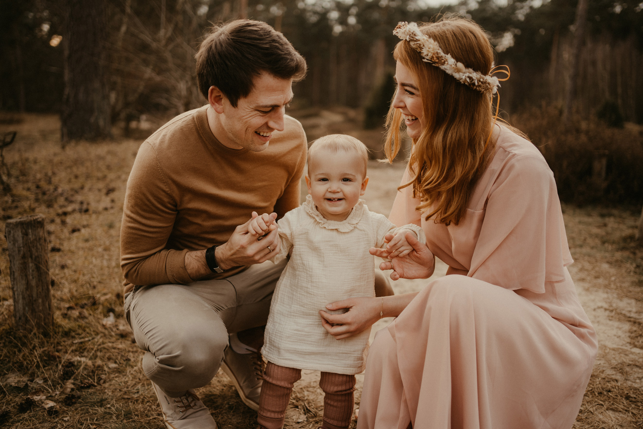 Eine Familie mit einem kleinen Kind steht im Wald während des Sonnenuntergangs in Münster. Die Eltern halten das Kind an den Händen und die Mutter trägt einen Blumenkranz auf dem Kopf.